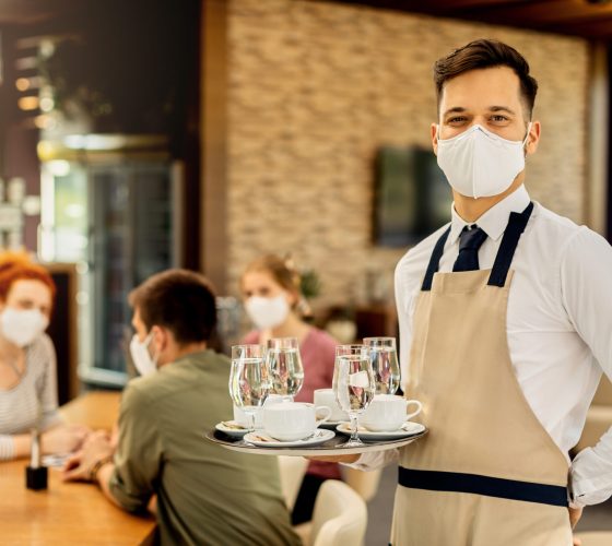 Portrait of happy waiter with protective face mask serving customers in a cafe and looking at camera.